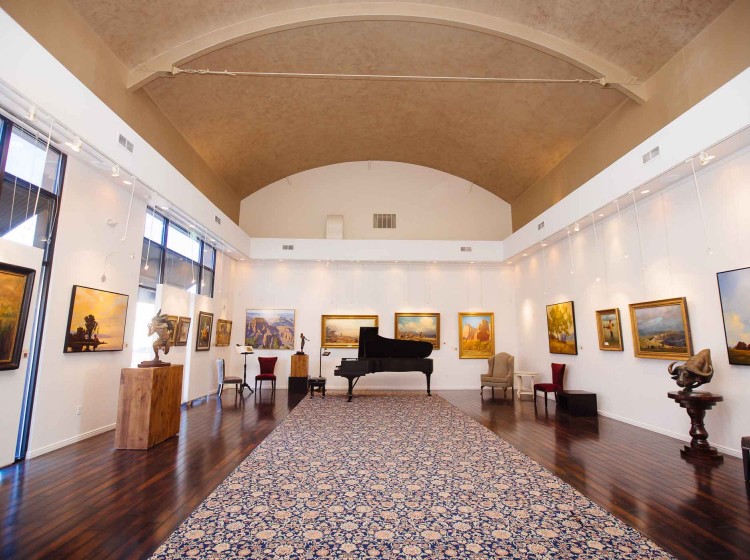 Interior of a bright gallery hall with a vaulted ceiling, patterned rug, grand piano, and framed artworks on white walls, calm and welcoming mood